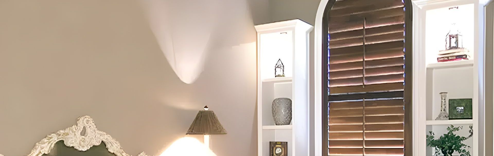 A bedroom with a bed , lamp , bookshelf and wood shutters.