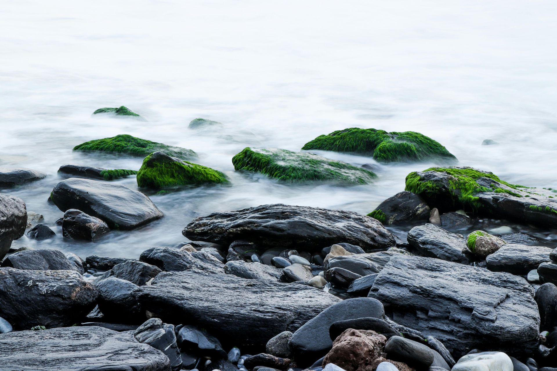 Rocks along a shore, some covered in green moss, with blurred white water and cloudy sky.