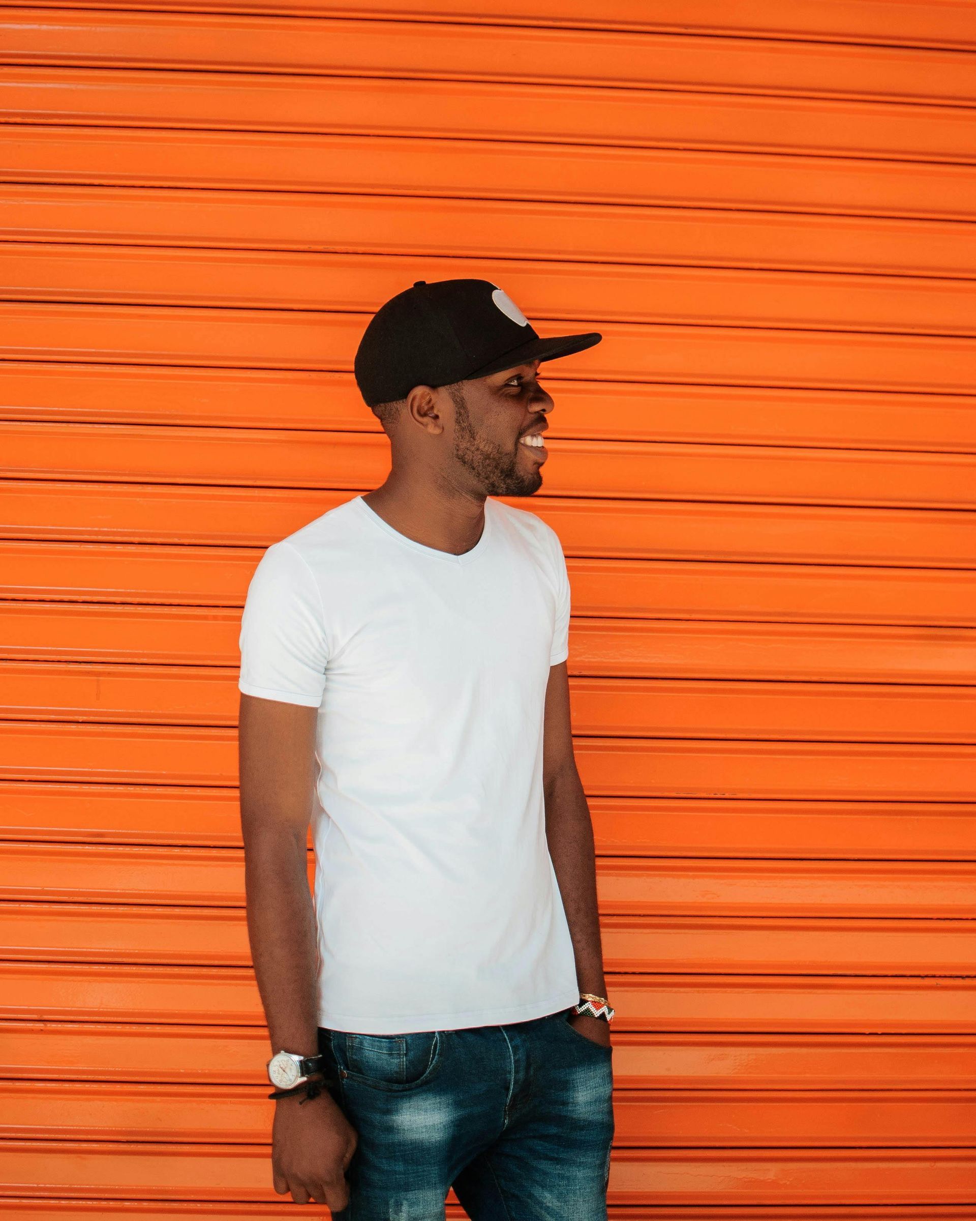 A man wearing a white t-shirt and a black hat is standing in front of an orange wall.