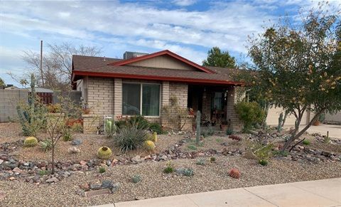 A single-story tan brick house with a brown roof and red trim, featuring a desert-style landscape with cacti and rocks.