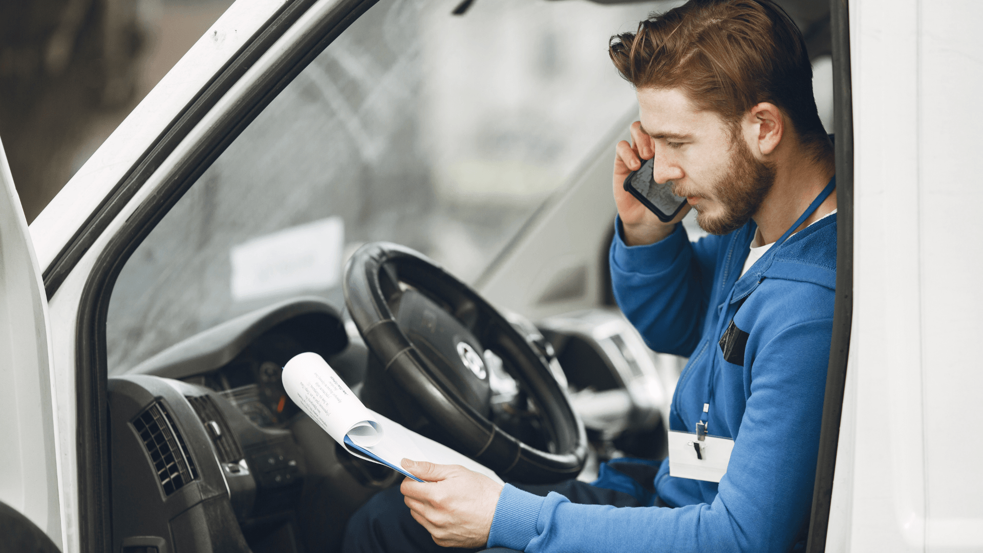 Man in blue uniform on phone in white van, reviewing paperwork.