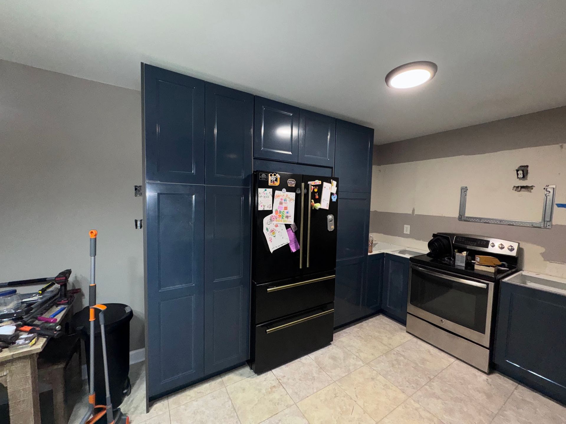 A kitchen with blue cabinets , a black refrigerator , a stove , and a trash can.