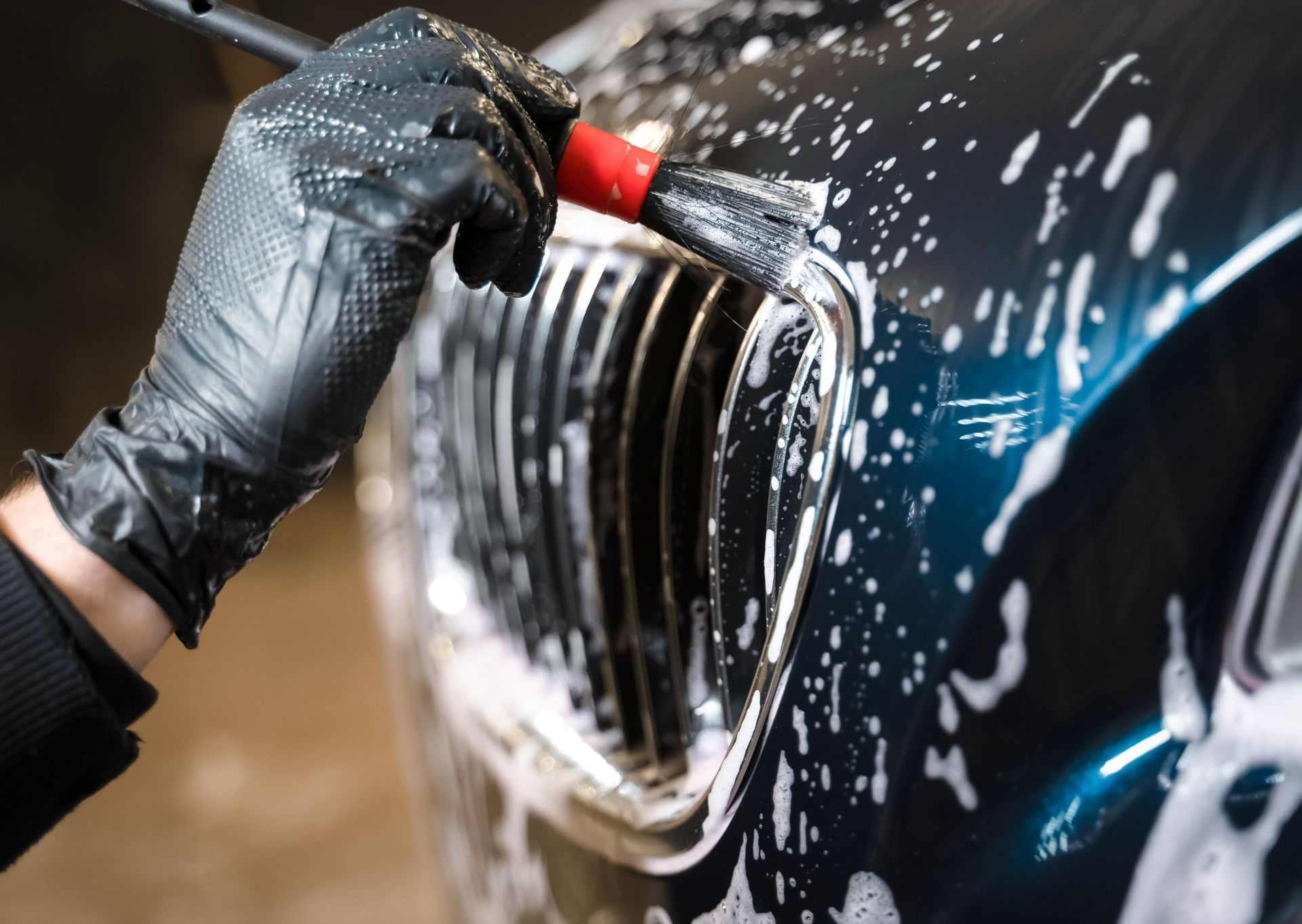 Black-gloved hand detailing a car's front grill with a brush and soapy water.