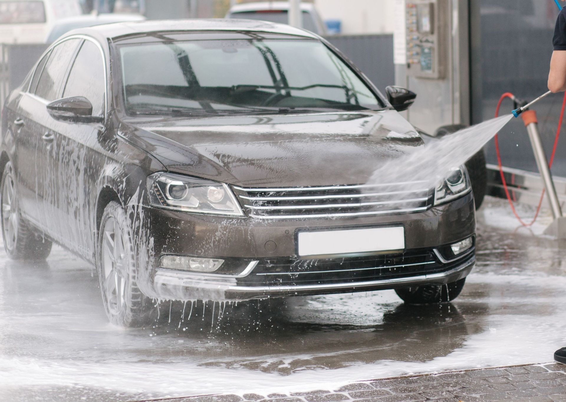 A dark sedan being washed at a car wash, water spraying from a hose, gray concrete floor.