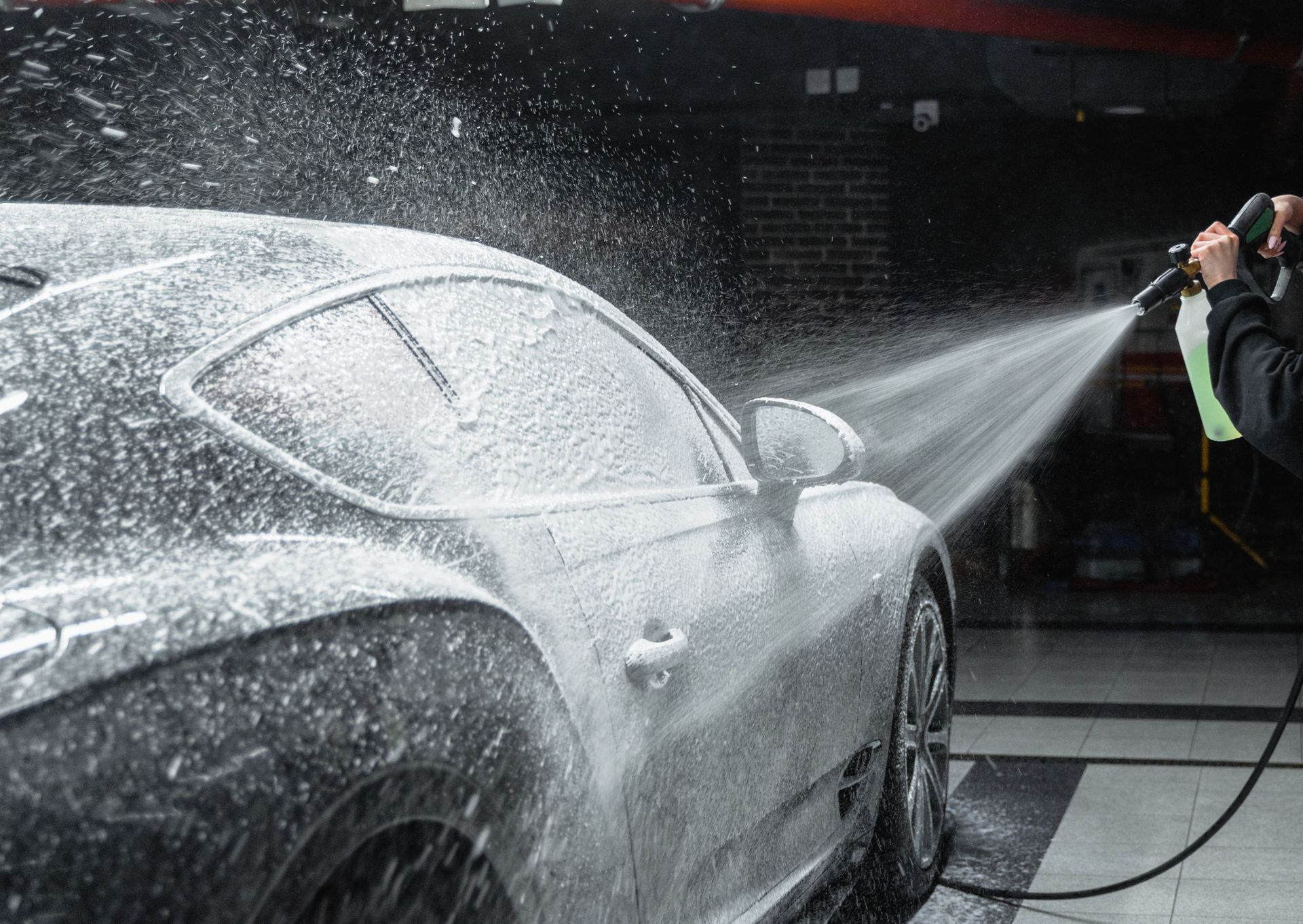 A person spraying foamy car wash solution on a sleek silver car in a garage.