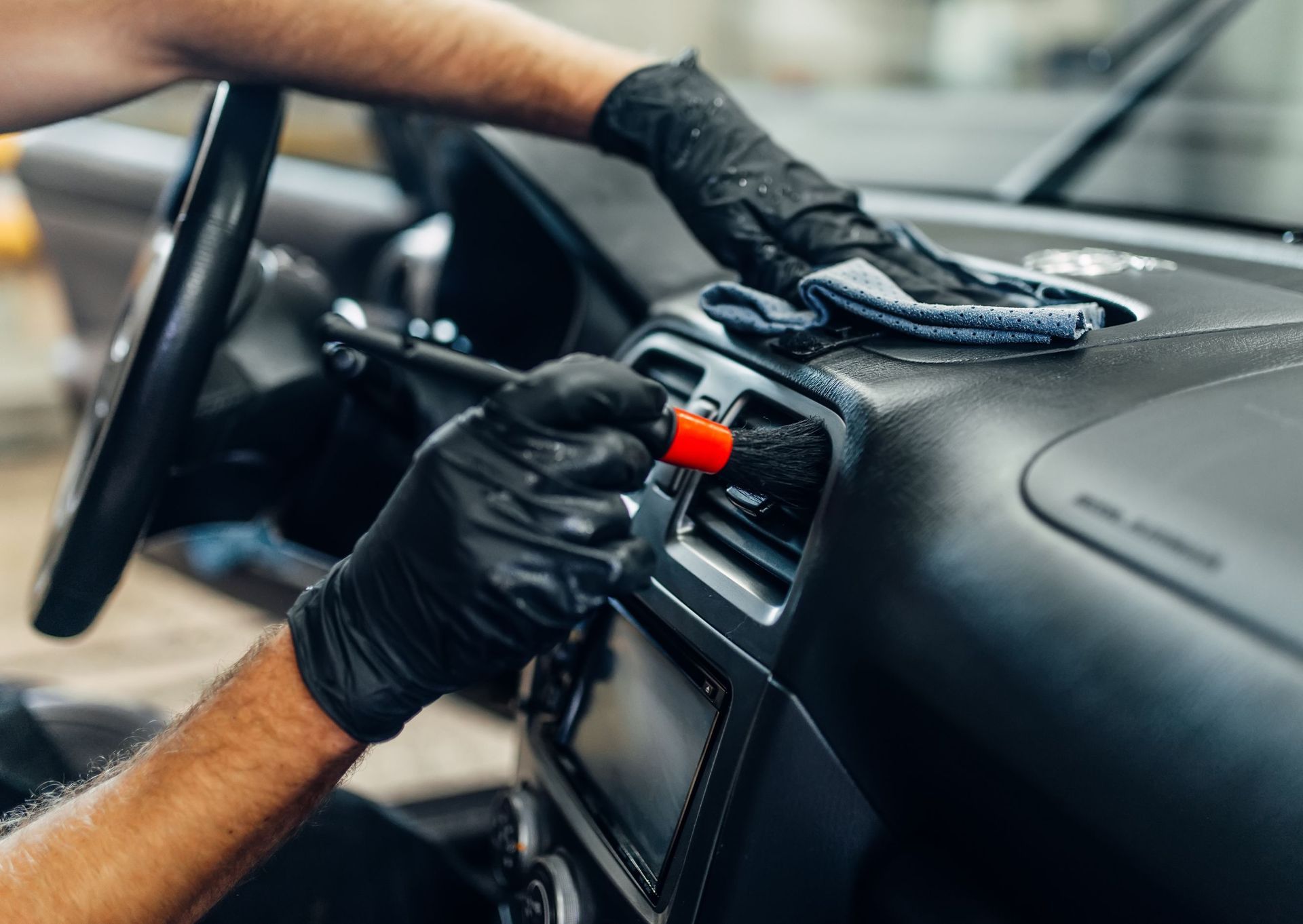 Person wearing black gloves cleaning a car dashboard with a brush and cloth.