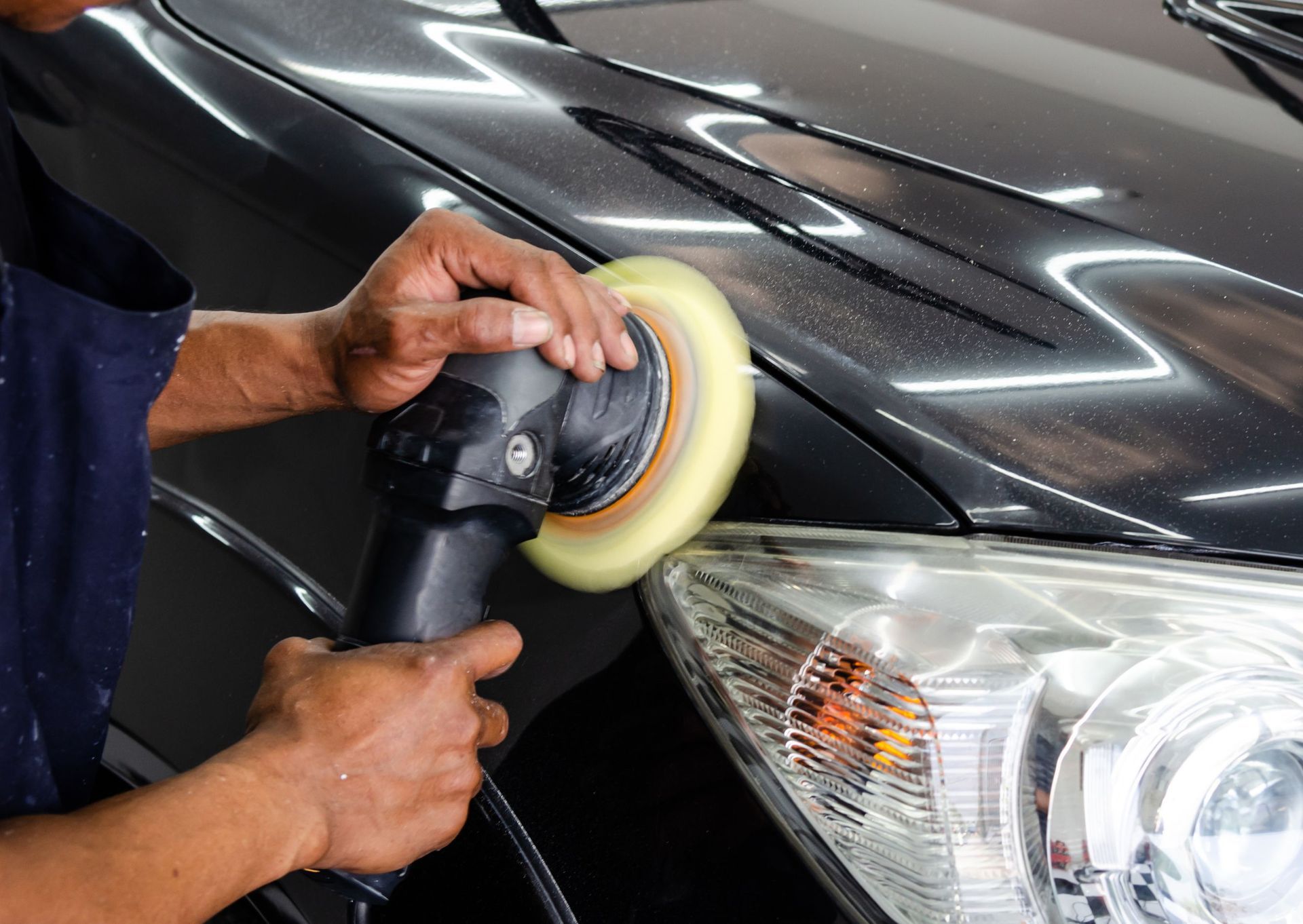 Person polishing a black car's hood with a buffer near a headlight.