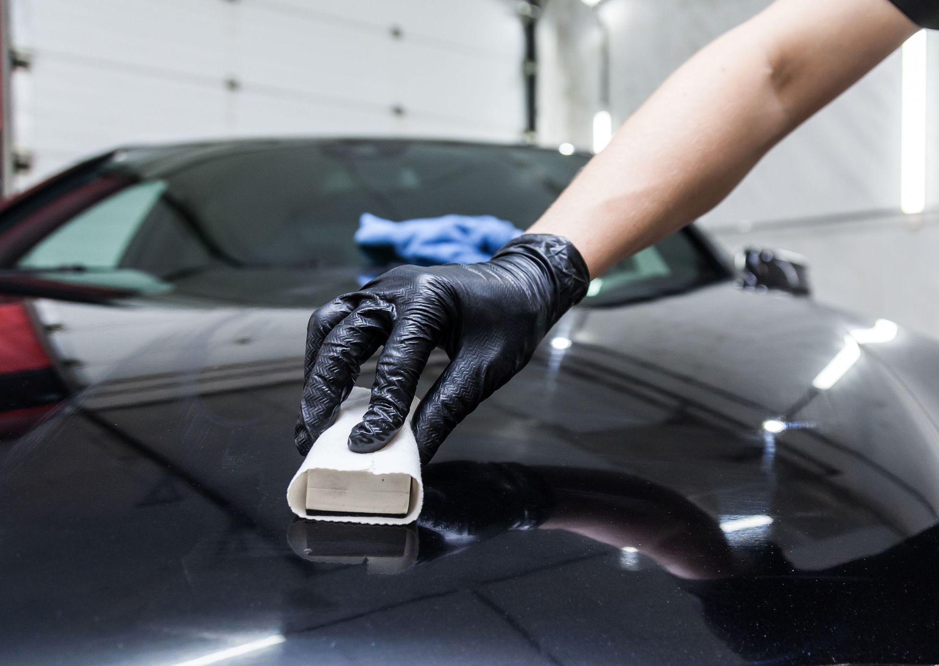 Person in black gloves applying ceramic coating to a black car hood in a garage.
