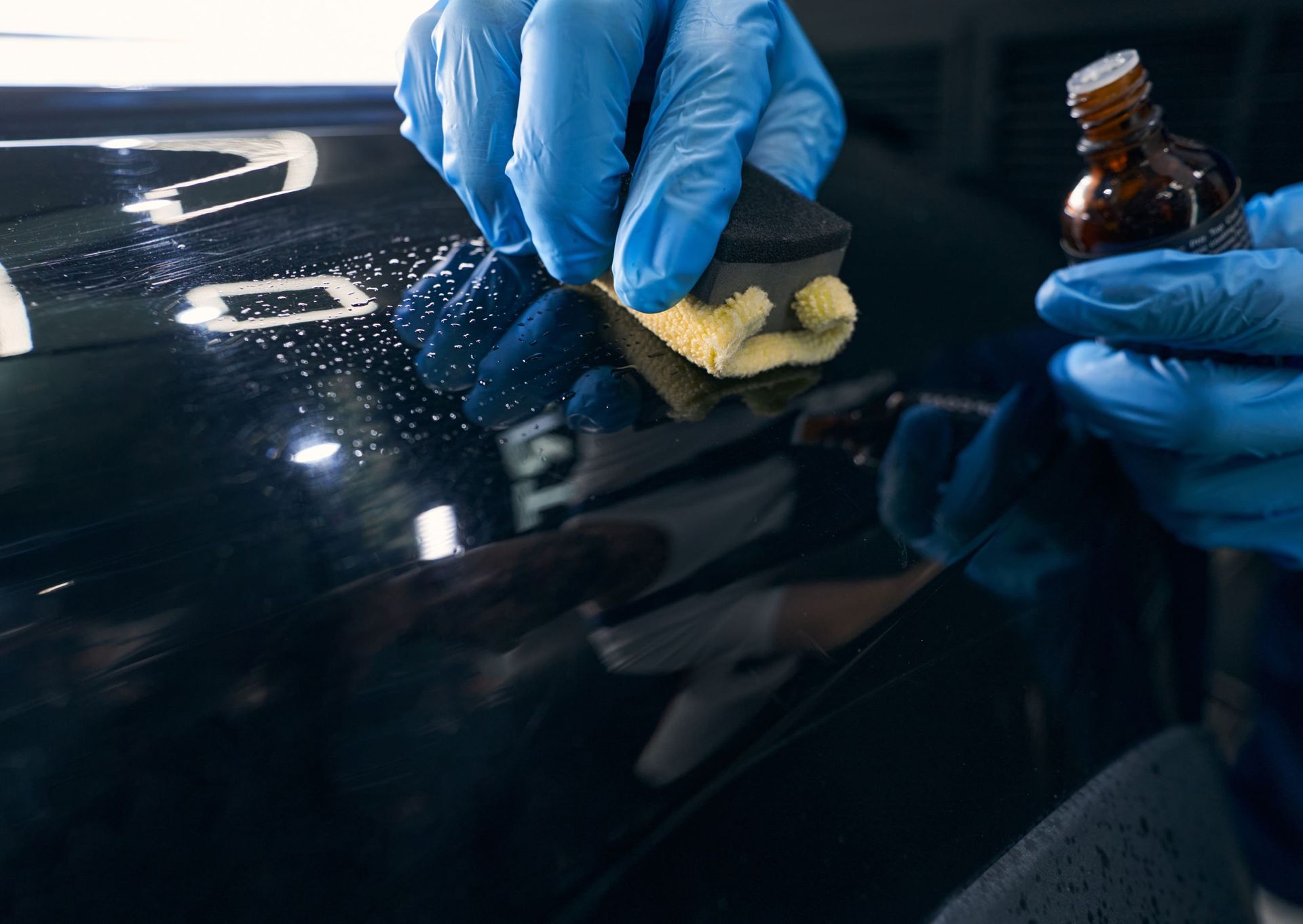 Hands in blue gloves applying ceramic coating to a car with a sponge and dropper bottle.