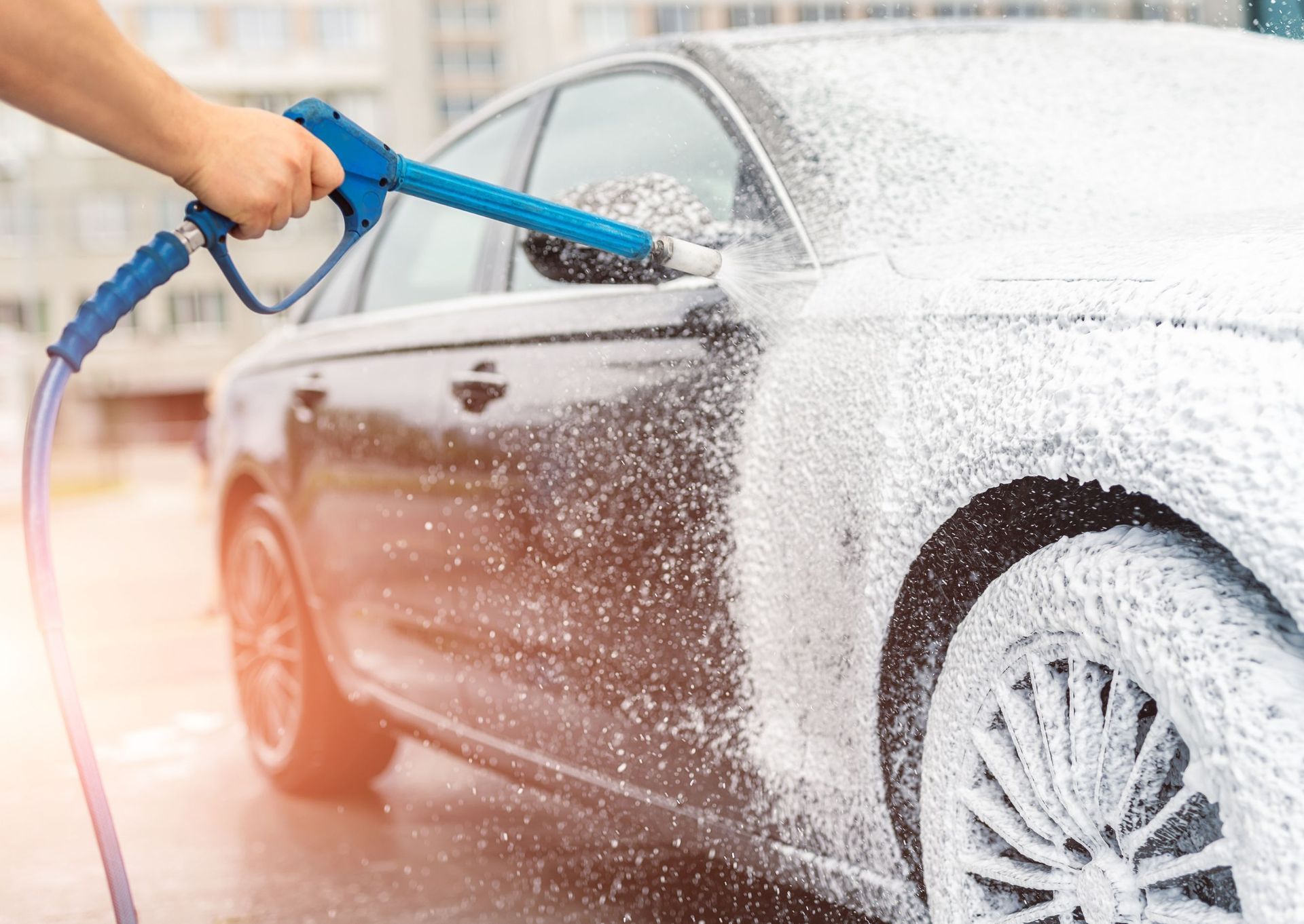 Person washing a dark car with foamy soap, using a blue spray nozzle.