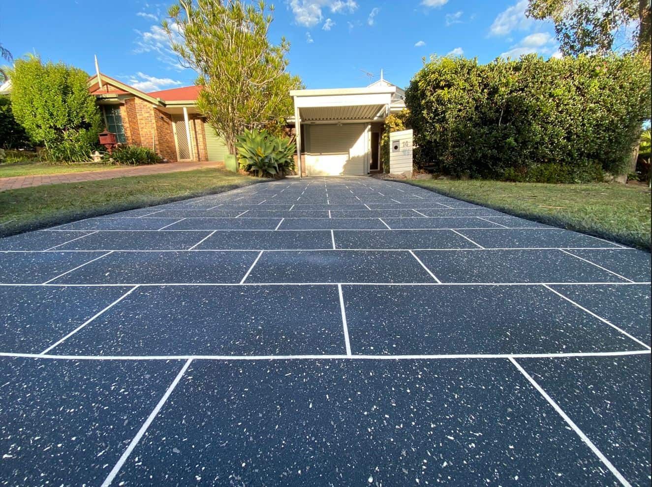 A low-angle view of a dark blue, speckled driveway with a rectangular tile pattern leading to a house and carport.