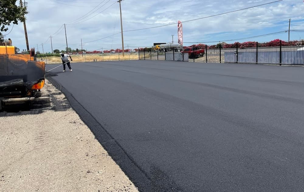 A construction worker smooths freshly laid black asphalt on a sunny day at an outdoor site.