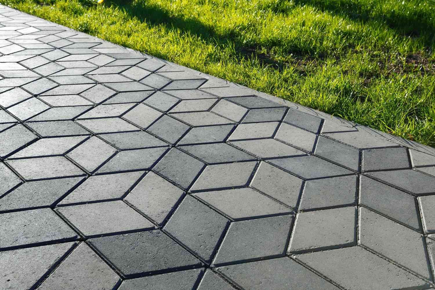 A paved walkway featuring gray diamond-shaped pavers in a 3D cube pattern, bordered by green grass.