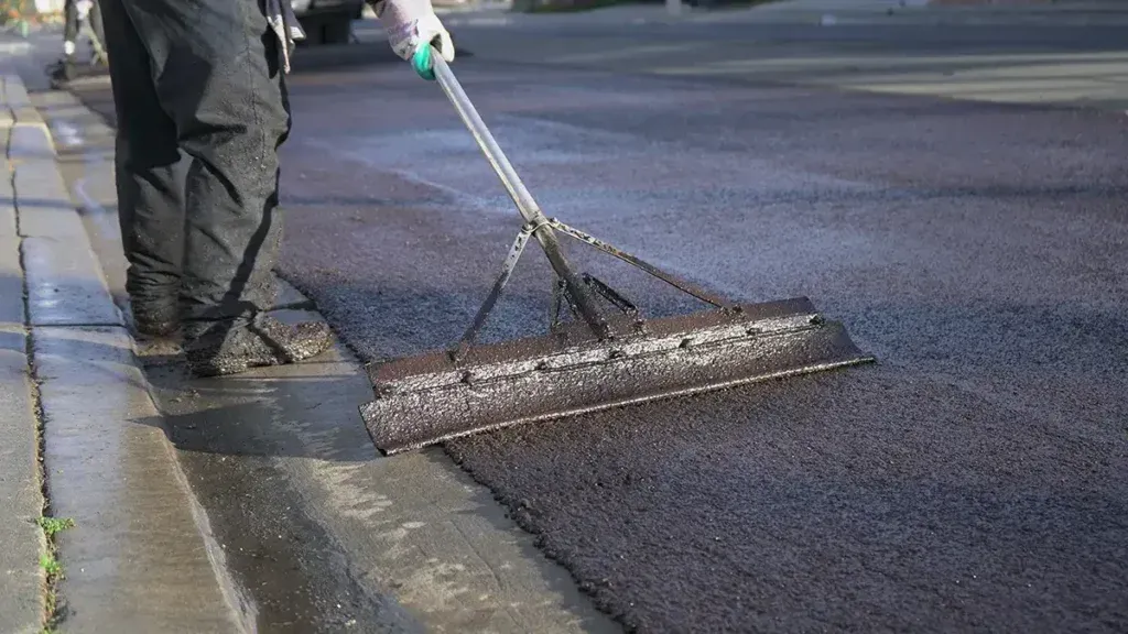 A worker uses a squeegee to spread fresh, black asphalt over a road surface.