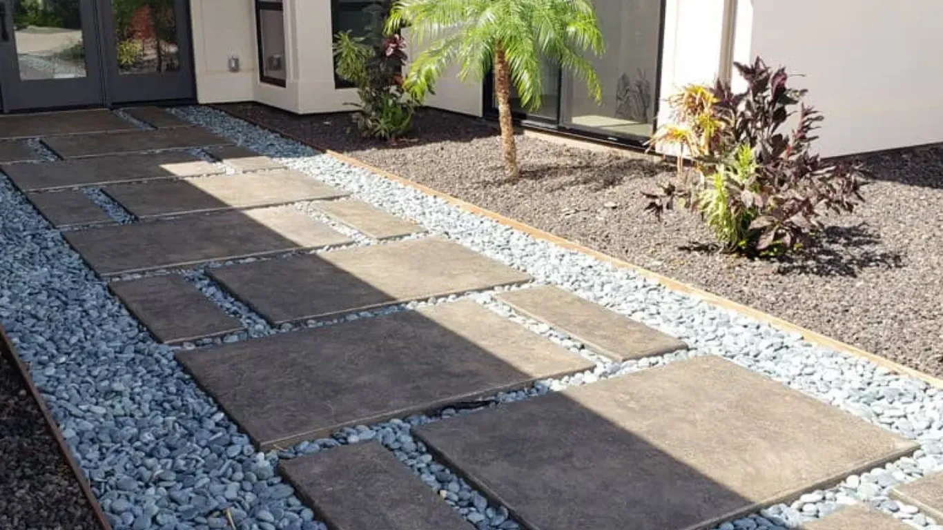 A modern stone paver walkway surrounded by grey gravel, leading to a home entrance with small ornamental plants.