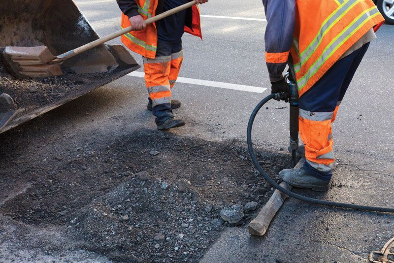 Two workers in high-visibility orange safety gear repair a pothole in an asphalt road using a jackhammer and a shovel.