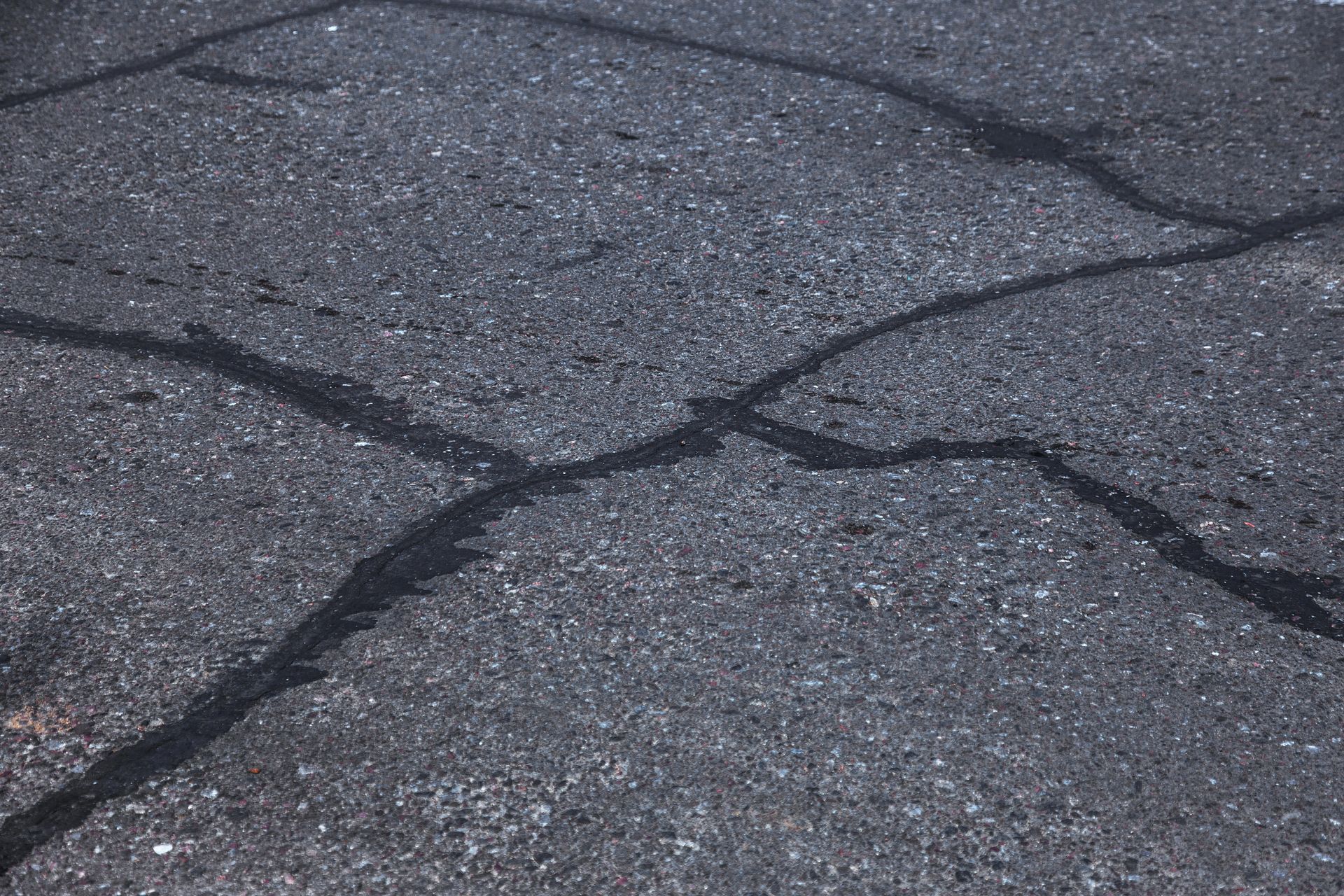 A close-up of dark grey asphalt pavement featuring a pattern of interconnected, deep cracks.