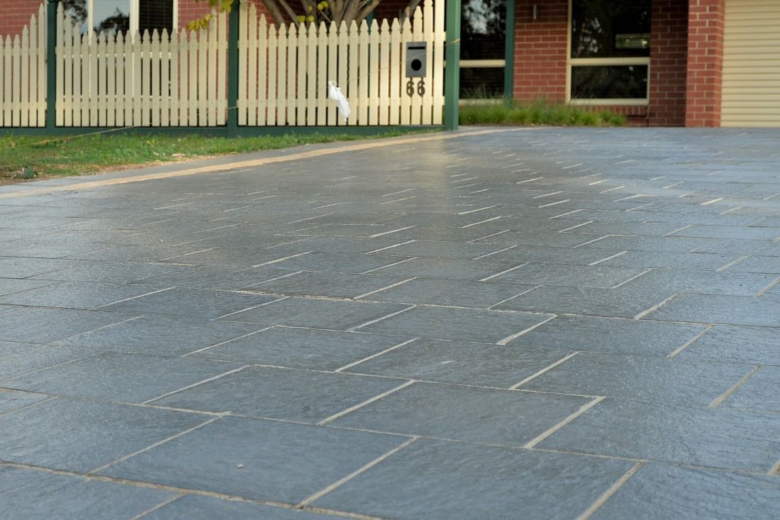 A textured, dark grey concrete driveway with a stamped, patterned design in front of a white picket fence and house.