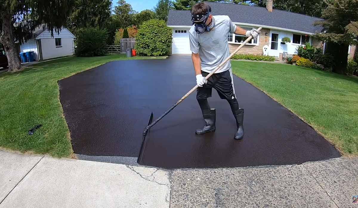 A person wearing protective gear spreads black asphalt sealant across a residential driveway with a long-handled brush.