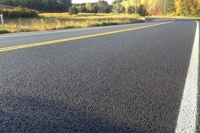 A freshly paved asphalt road curves through a rural landscape with yellow center lines and white side lane markings.