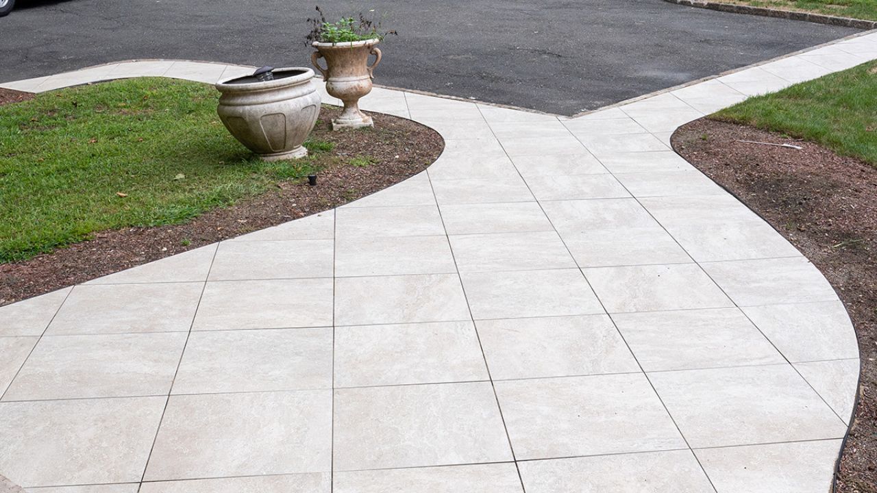 A paved walkway curves through a grassy garden area, leading toward a dark driveway with two decorative stone planters.