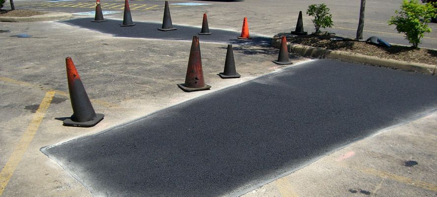 Freshly paved black asphalt patches on a weathered parking lot, marked off by several orange and black traffic cones.