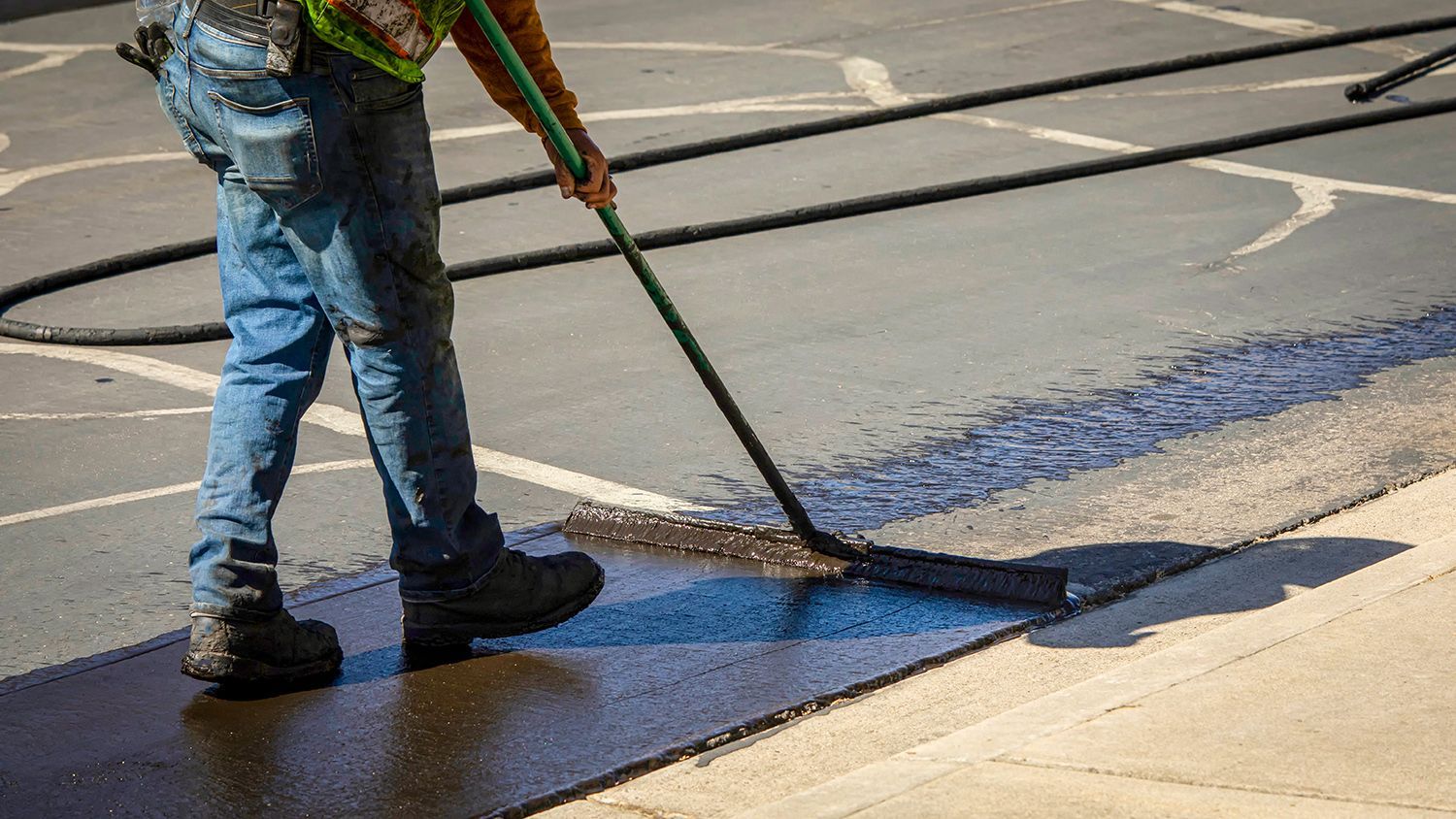 A worker in work clothes uses a long-handled squeegee to spread black asphalt sealant across a paved surface.