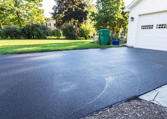 A newly paved, dark asphalt driveway leads to a white garage, flanked by a green lawn and trees in a residential setting.