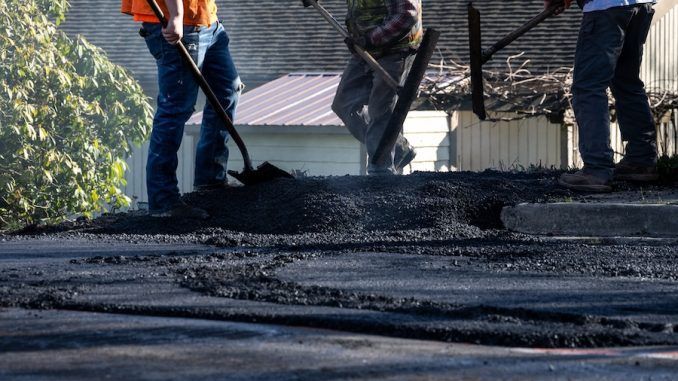 Construction workers spread fresh asphalt on a road surface using shovels and rakes.