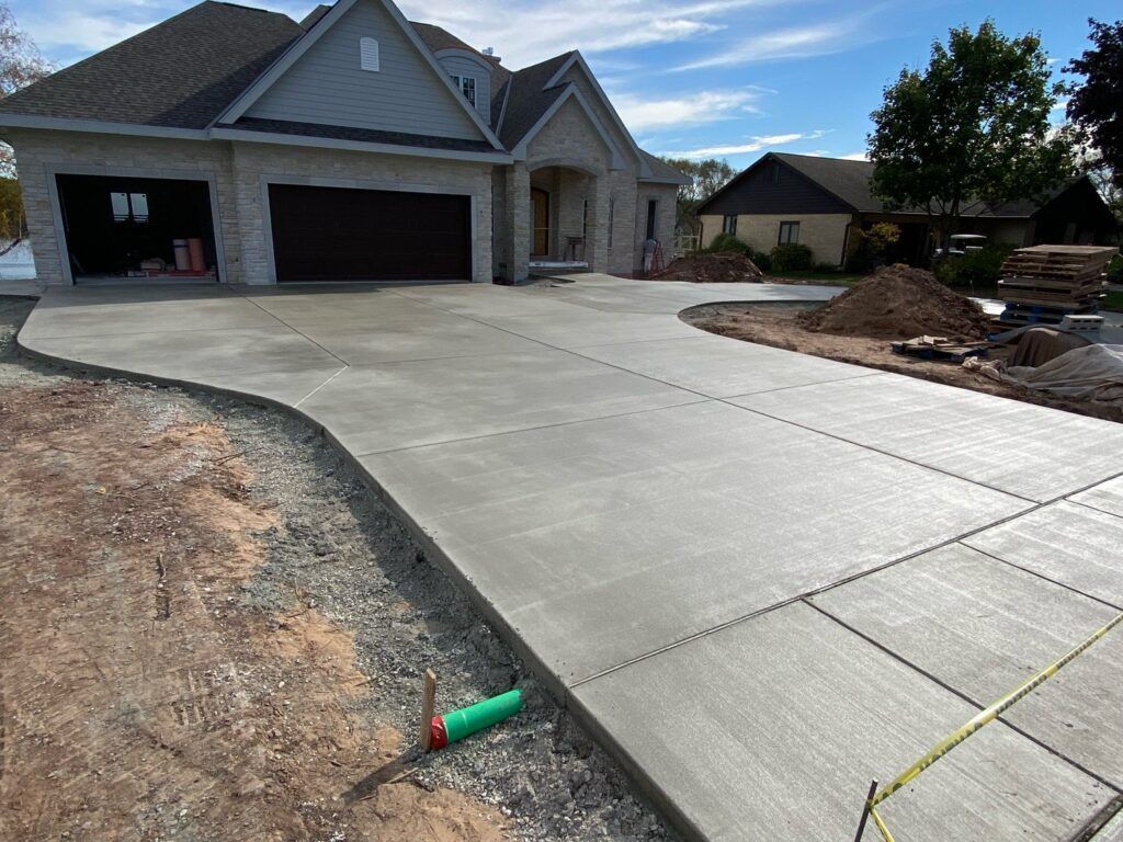 A newly poured concrete driveway leads to the garage of a light-stone house under a sunny blue sky.