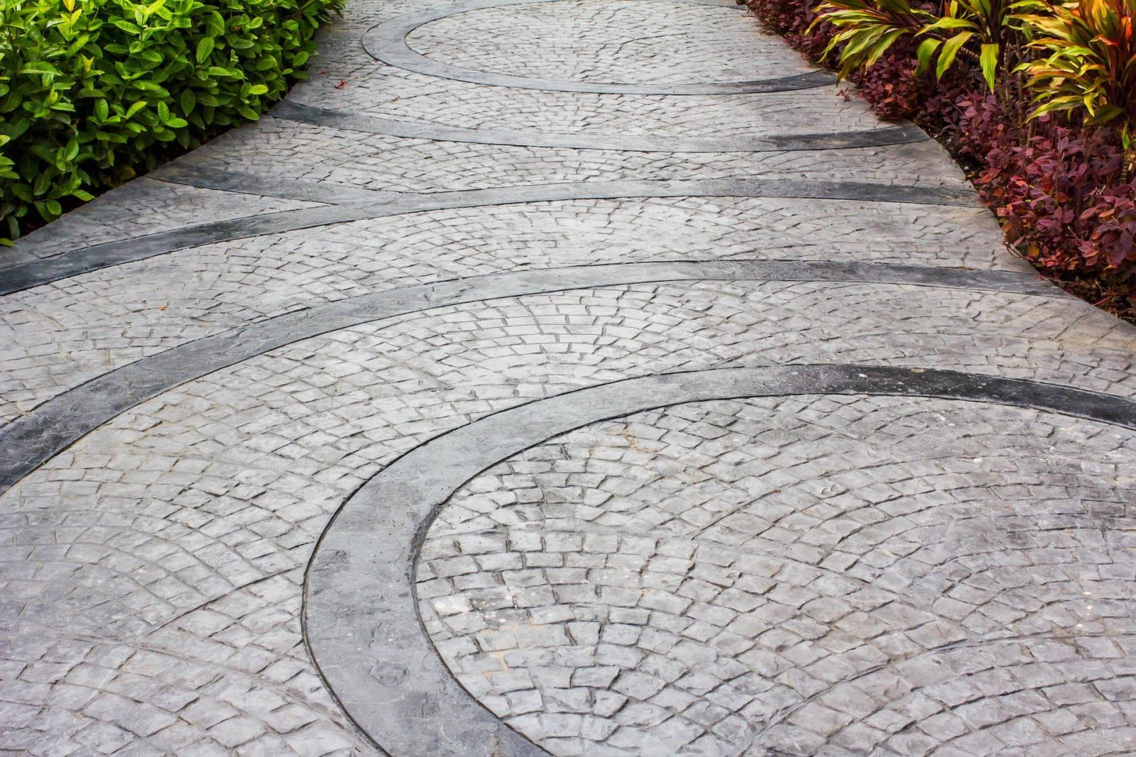 A stone walkway with a curved, patterned design, bordered by lush green bushes on one side and colorful plants on the other.