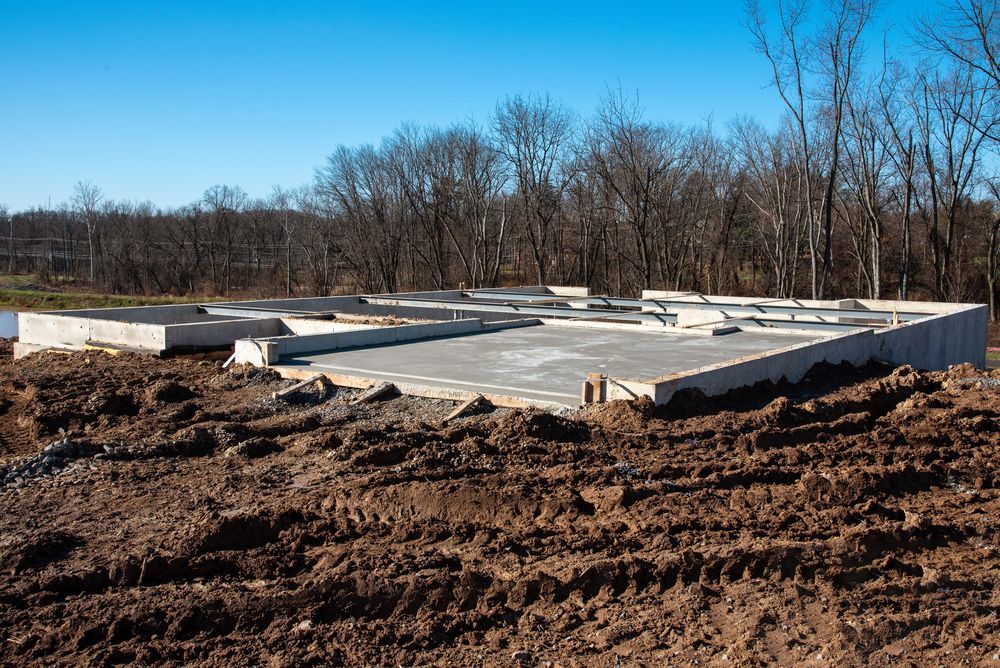 A concrete foundation for a building sits on a cleared, muddy construction site under a clear blue sky.