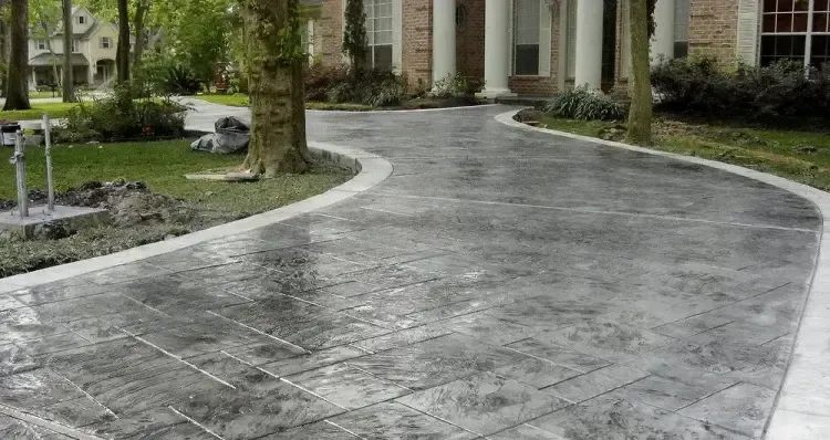 A gray, stamped concrete driveway curves toward the entrance of a brick house with white columns.