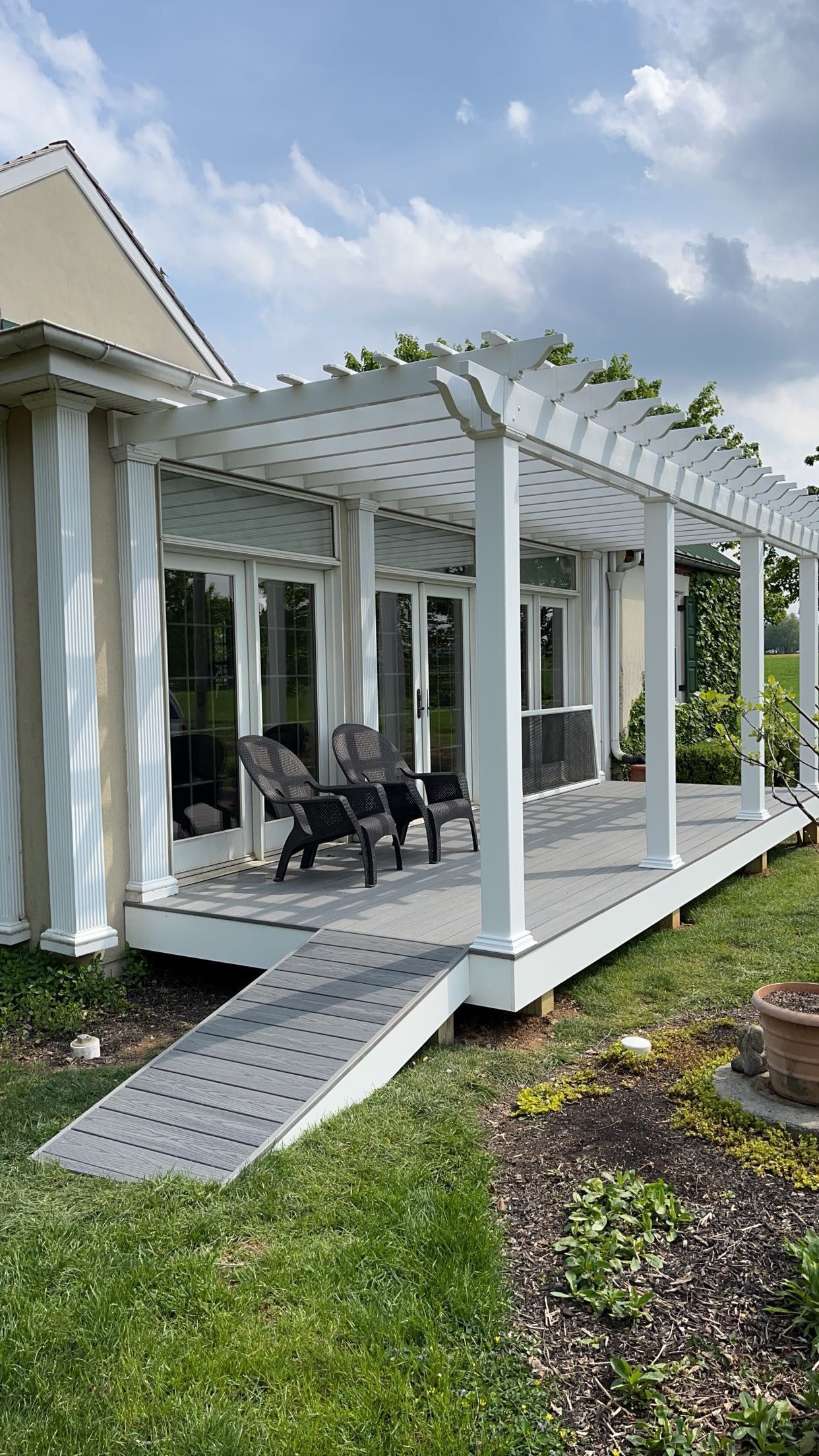 White pergola-covered patio with black chairs, ramp, and sliding glass doors beside a house