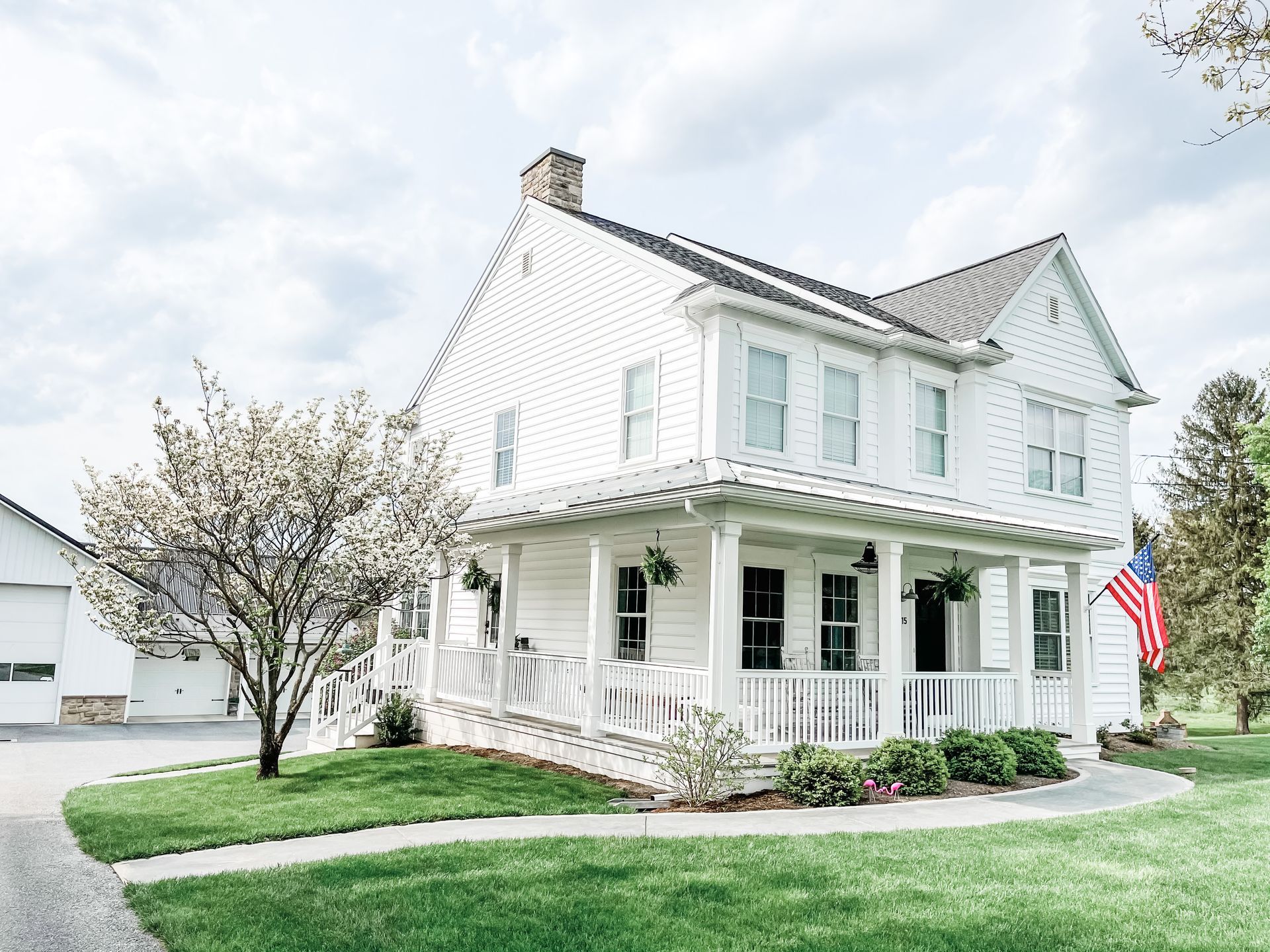 White two-story house with a front porch, blooming tree, and American flag on a green lawn