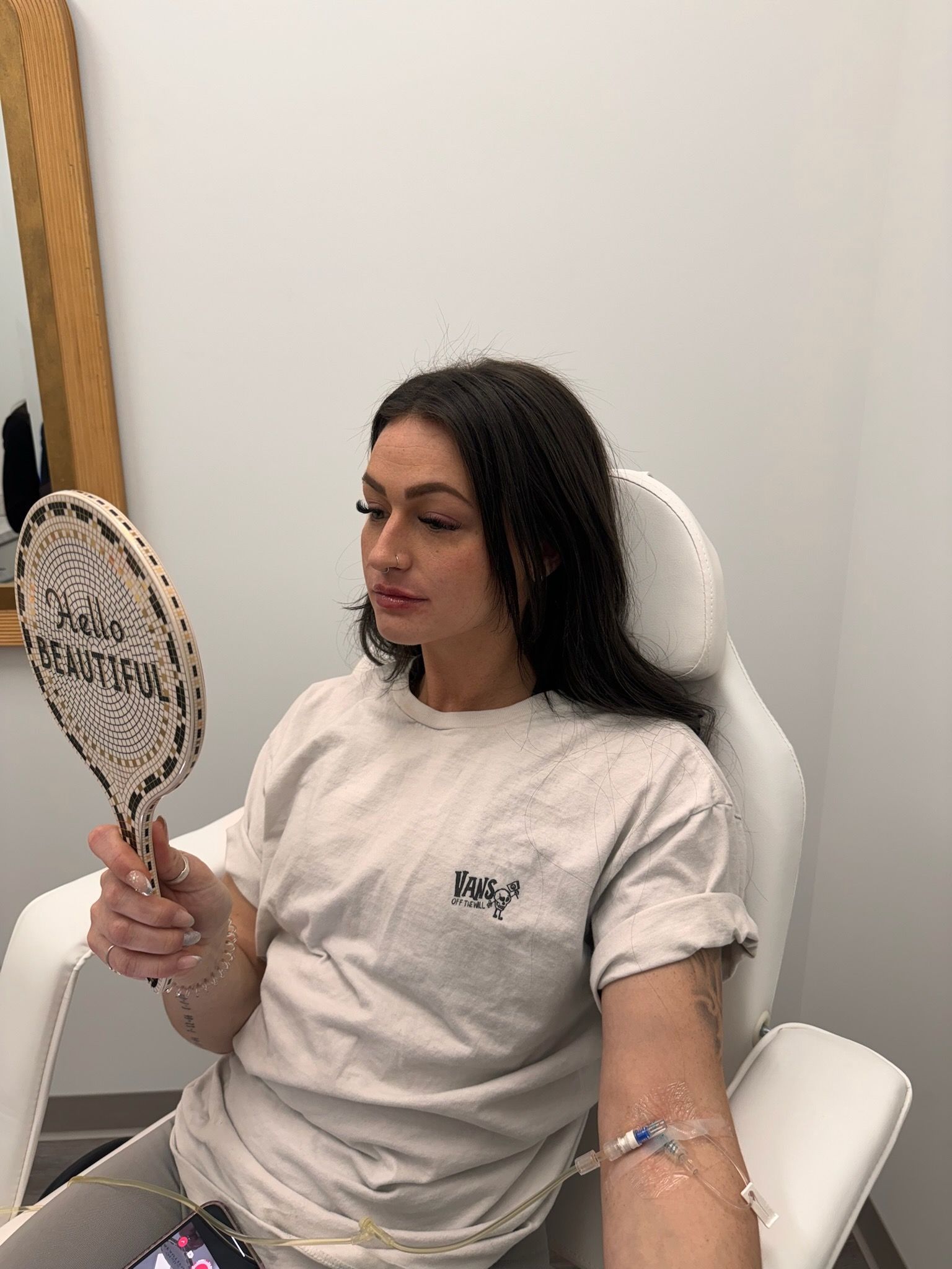 Woman in chair with IV, looking in hand mirror, possibly at face. Neutral-toned room.
