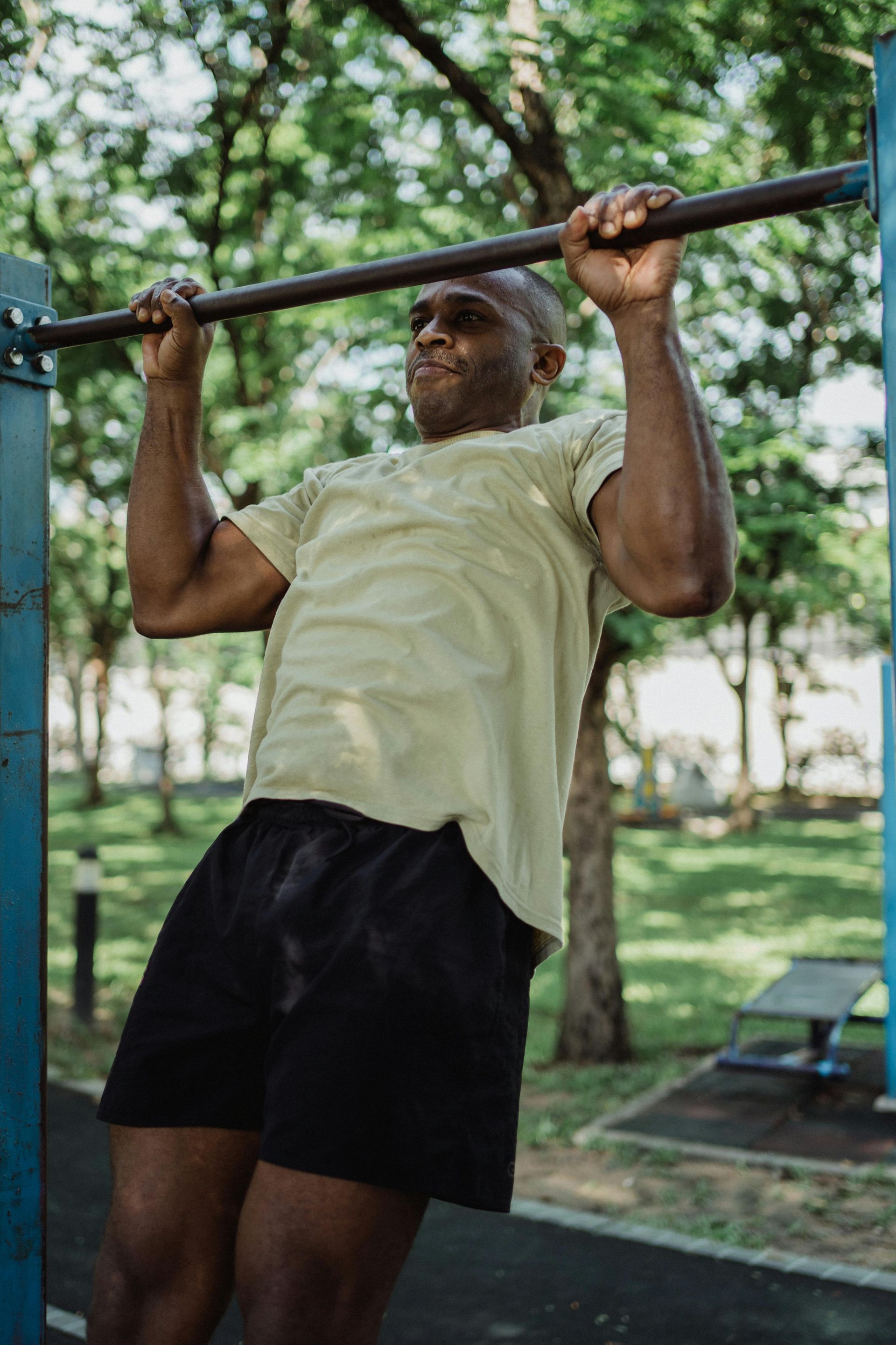 Man doing a pull-up on a bar outdoors. He wears a green shirt and black shorts in a park.