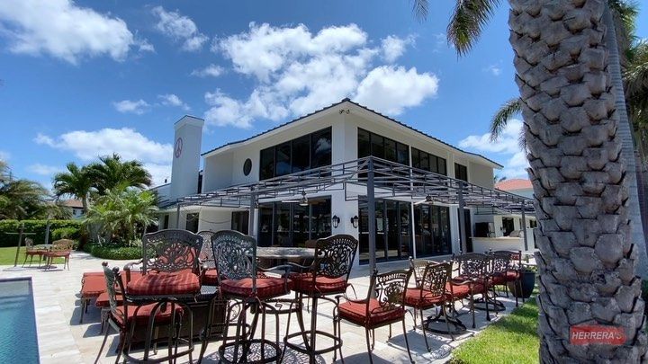 Sunny patio with red chairs outside a modern white house, framed by palm trees and blue sky.