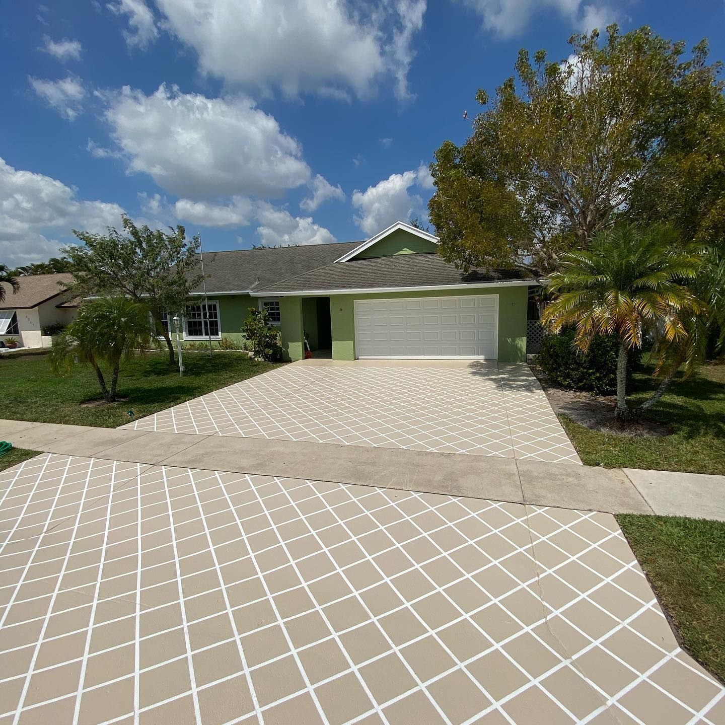 Suburban house with a wide paver driveway, two-car garage, green lawn, and a tree under a partly cloudy sky