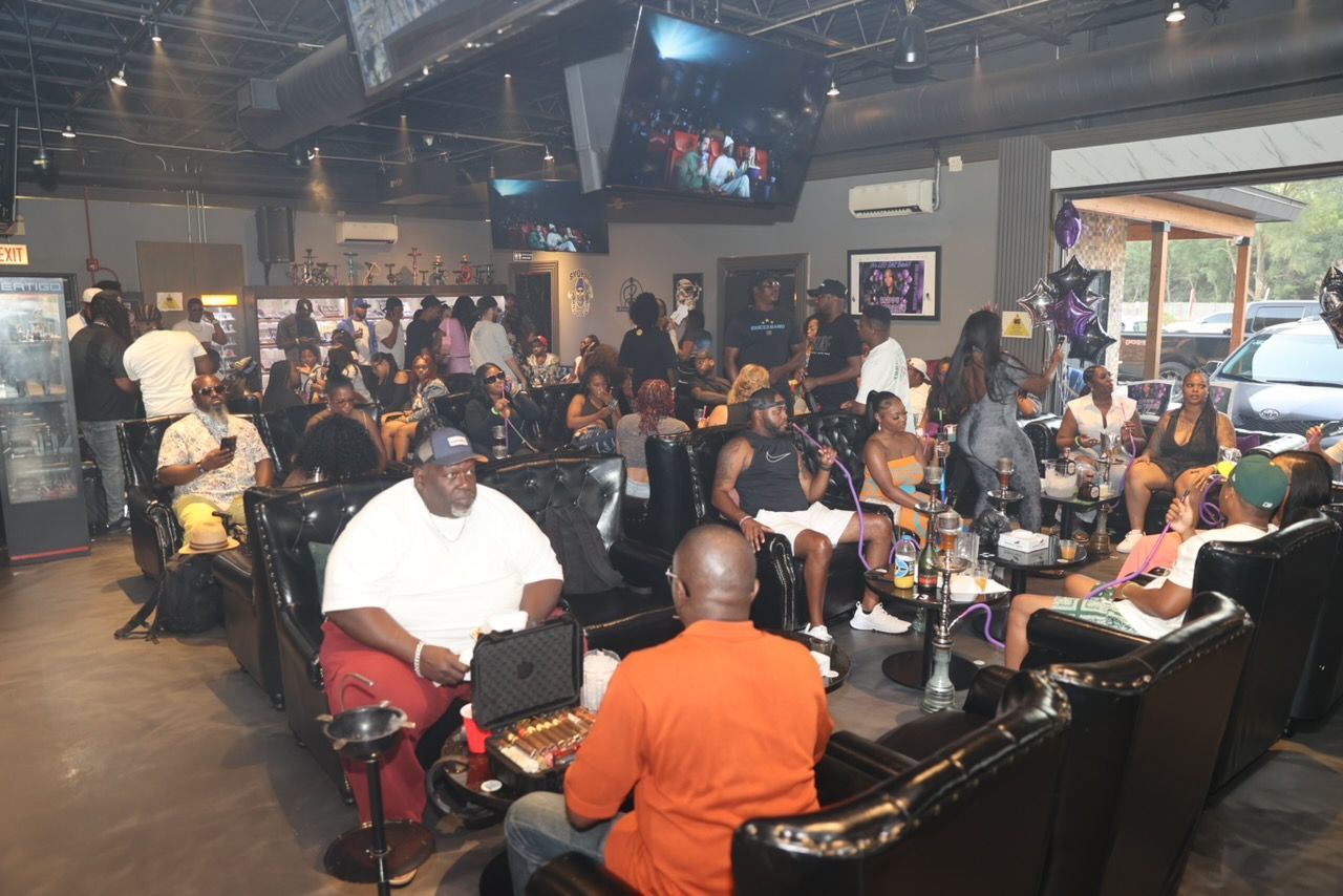 Large group of people gathered indoors at a hookah lounge, socializing and smoking.  Dark leather seating, TVs on walls, and low lighting.