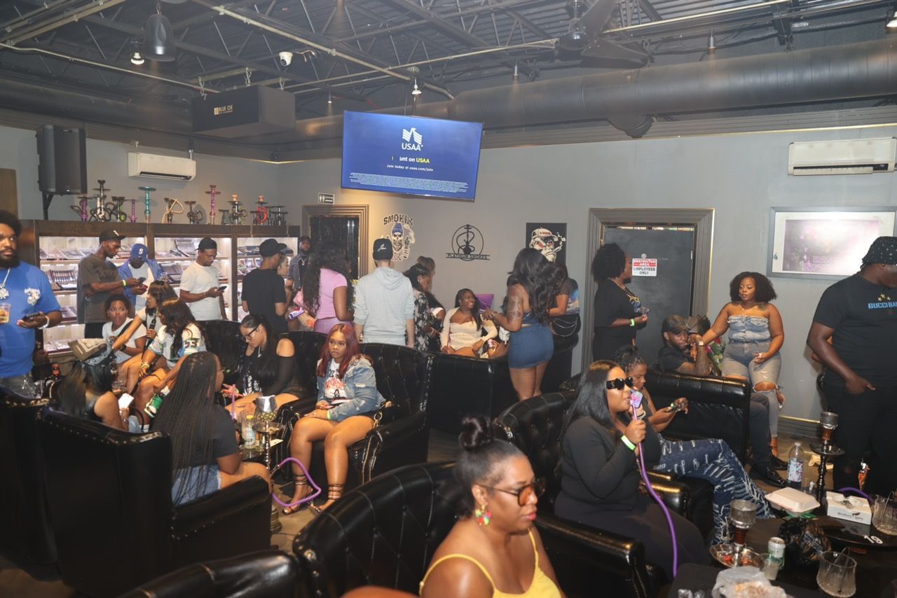 People gather in a dimly lit lounge. Some are seated, socializing, others standing near a bar. A TV screen is visible on the wall.