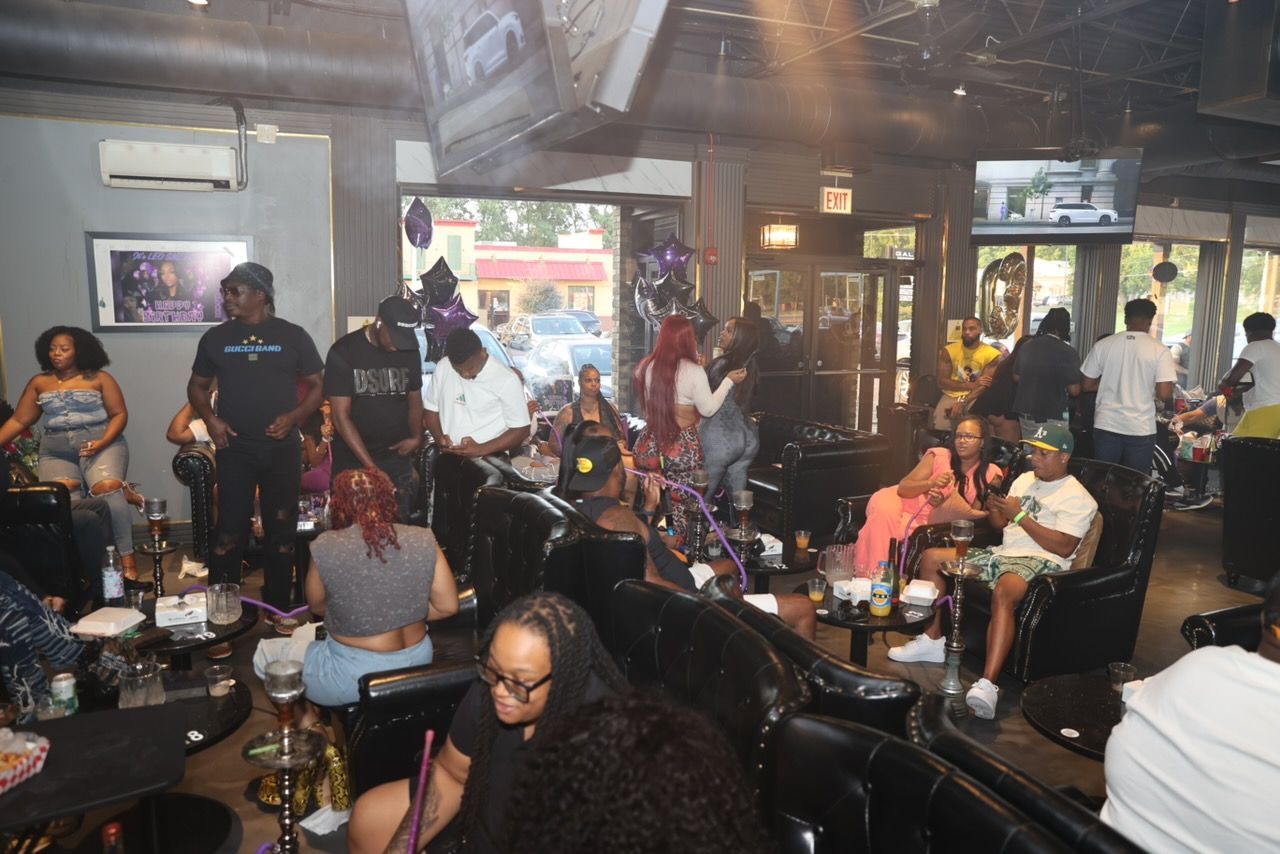 A crowded, dimly lit lounge with people socializing, some smoking hookahs. The space has black furniture and decorations with an open doorway.