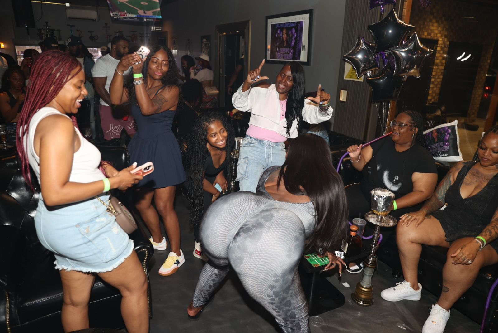 A group of Black women partying and dancing in a dim club. Some are smiling, while one in a grey jumpsuit twerks.