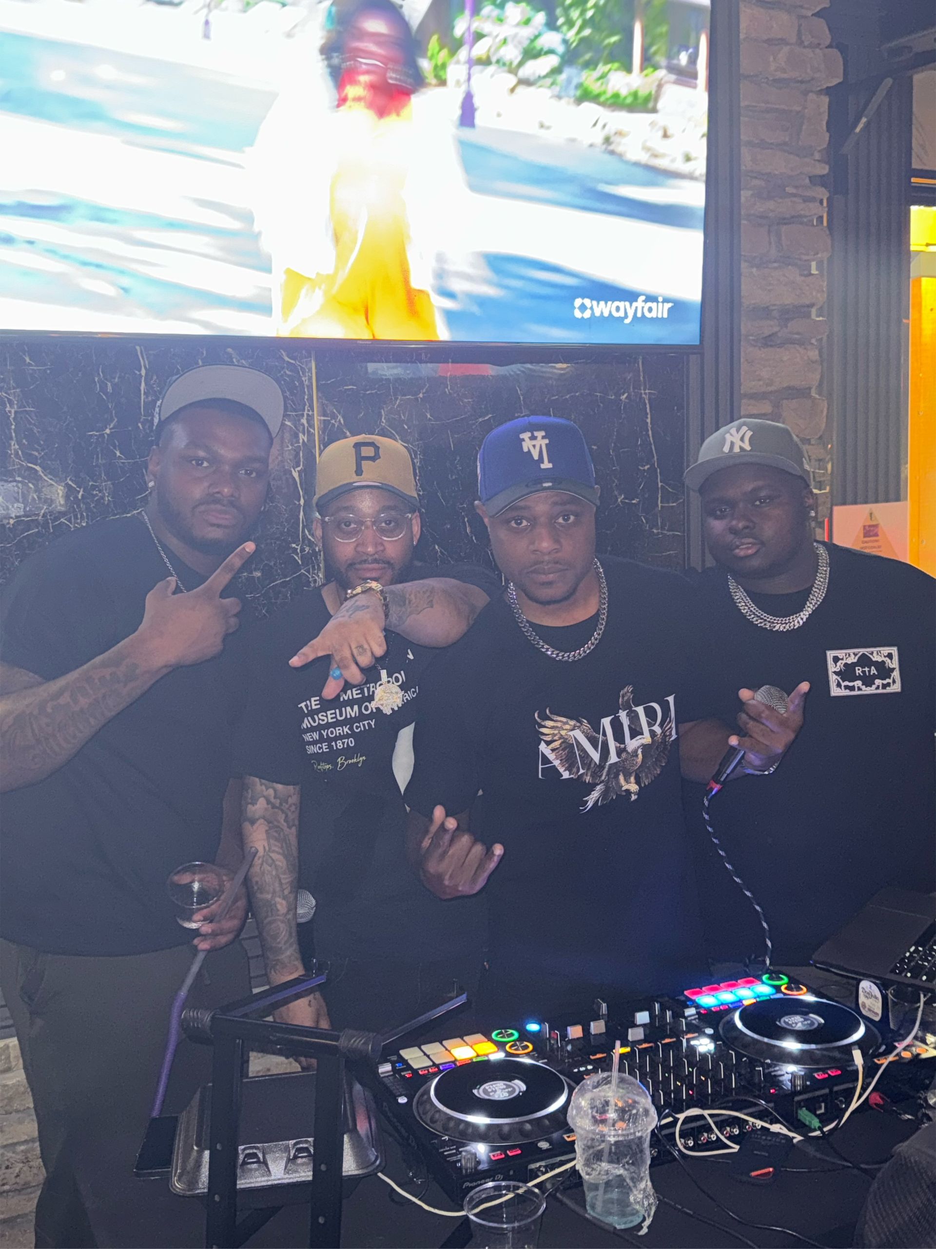 Four men pose with a DJ setup. They wear casual clothing and hats, with one pointing and the others gesturing. The setting appears to be an outdoor event.