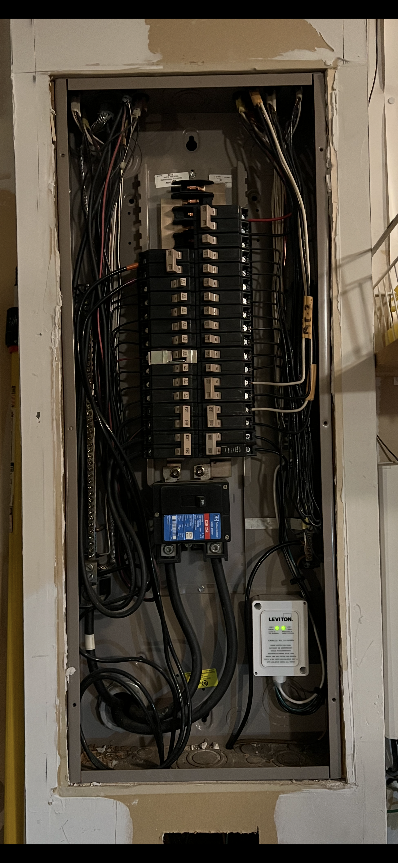 Electrical panel with numerous wires and circuit breakers inside a wall opening.