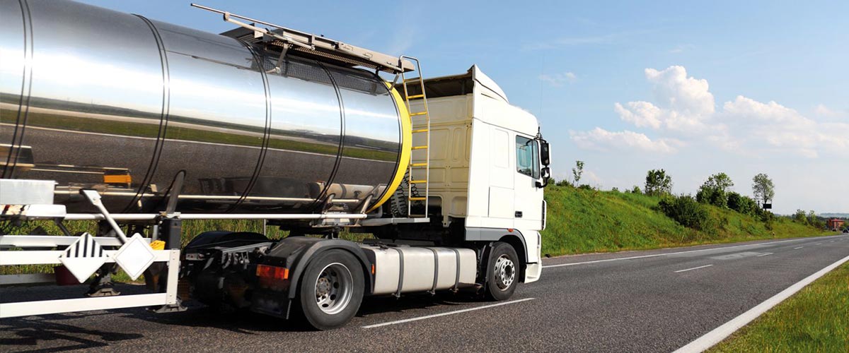Tanker truck driving on a highway, sunny day, green hill on the right.