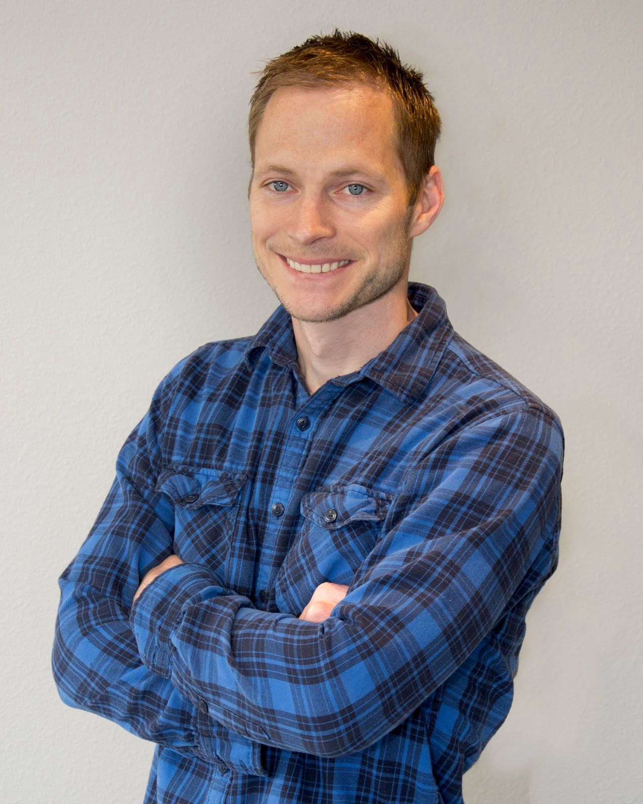 A man in a blue plaid shirt is standing with his arms crossed and smiling.