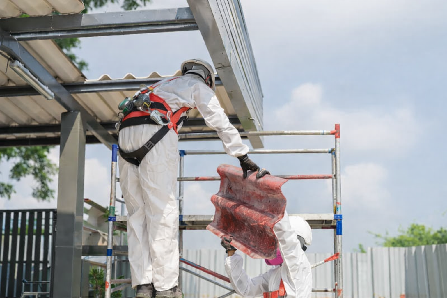 Due operai in tuta protettiva rimuovono le tegole. Struttura metallica, giornata di sole.