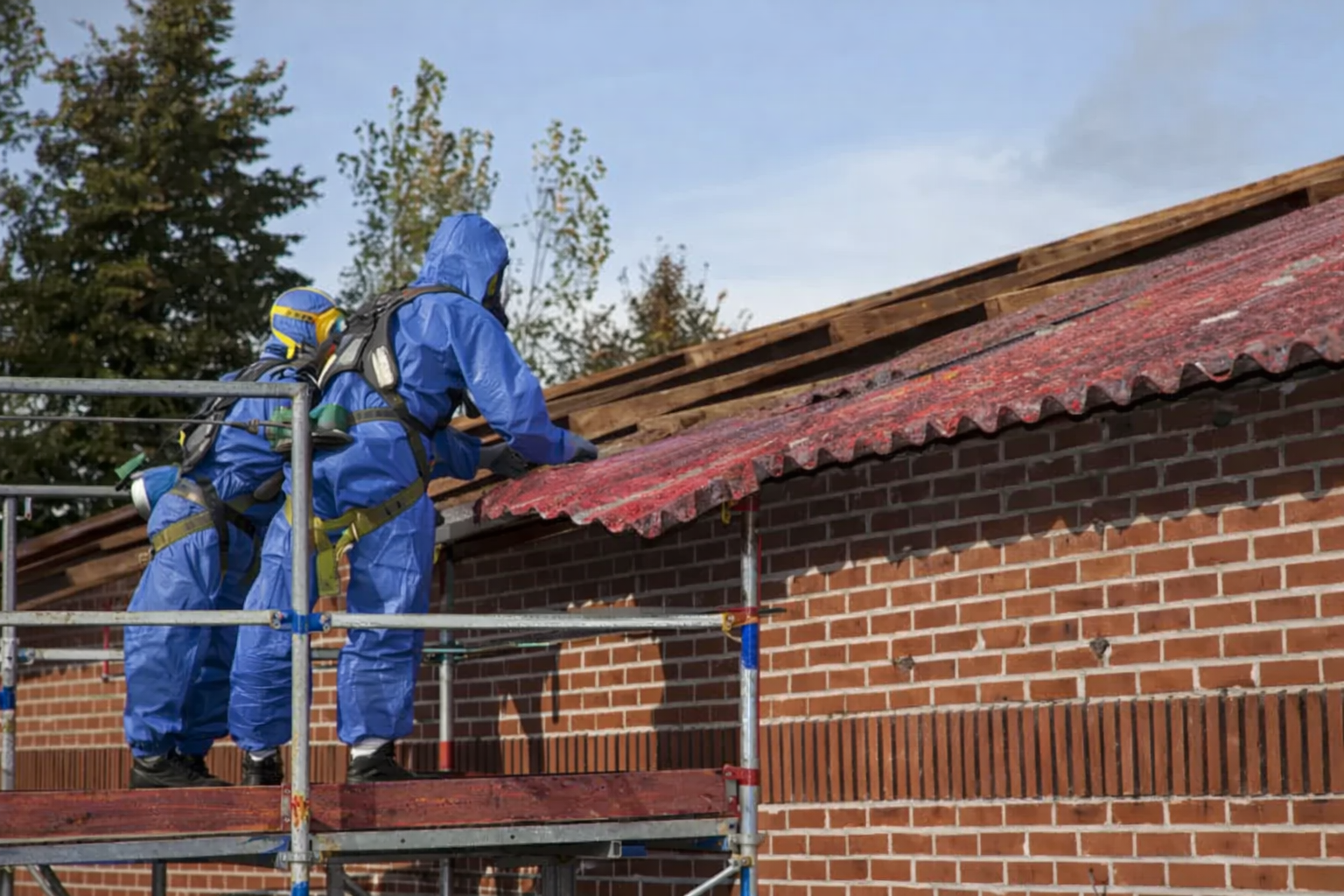 Due operai in tute protettive blu su un ascensore stanno rimuovendo un tetto ondulato da un edificio in mattoni.