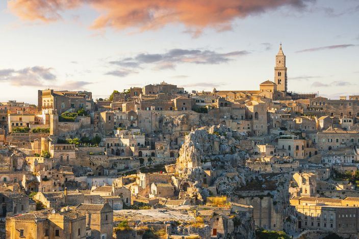 Paesaggio urbano di Matera, Italia, con antichi edifici in pietra e un campanile sotto un cielo nuvoloso al tramonto.